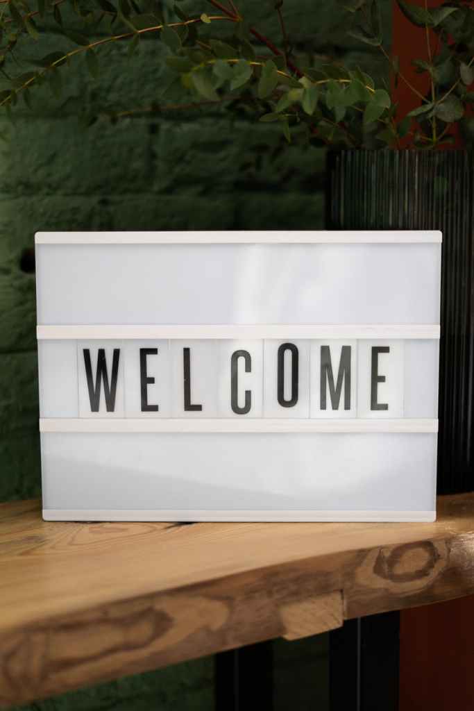 Image description: a white lightbox sign with the word WELCOME sits on top of a wood table. Faintly behind the sign is a wall with bricks and a plant.